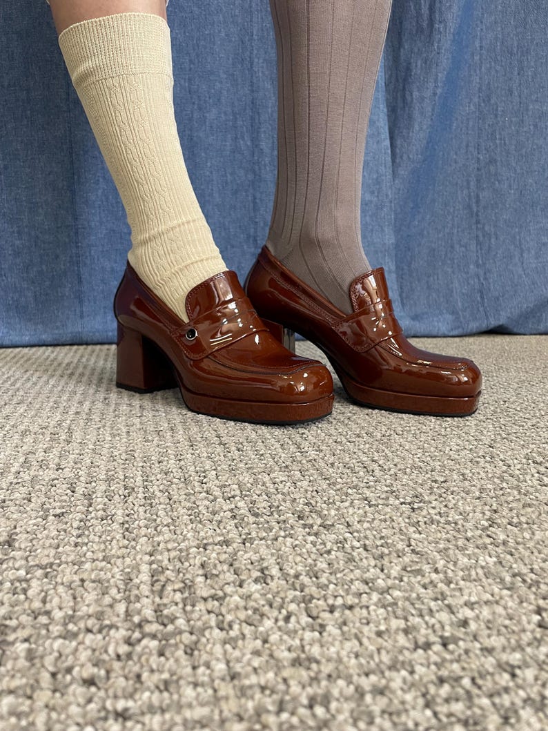 May include: Brown patent leather platform loafers with a chunky heel. The shoes have a classic penny loafer design with a rounded toe and a small metal detail. The image shows the shoes from the ankles down, with beige and brown socks.