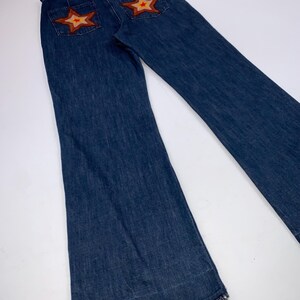 May include: Dark blue denim jeans with flared legs. The back pockets feature star-shaped patches with orange and brown accents. The jeans have a classic five-pocket design and a frayed hem.