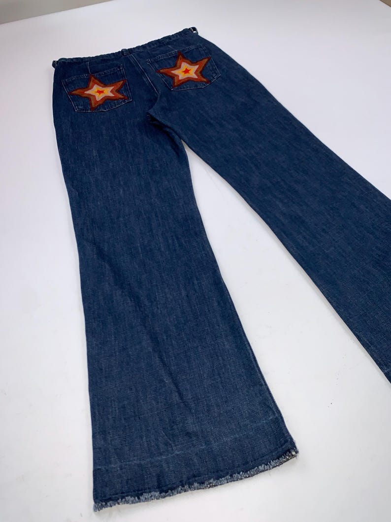 May include: Dark blue denim jeans with flared legs. The back pockets feature star-shaped patches with orange and brown accents. The jeans have a classic five-pocket design and a frayed hem.
