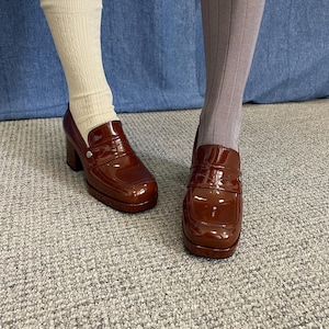 May include: Pair of brown patent leather platform loafers. The shoes have a square toe, a chunky heel, and a decorative strap across the vamp. One foot is wearing a cream-colored sock, and the other is wearing a gray ribbed stocking.