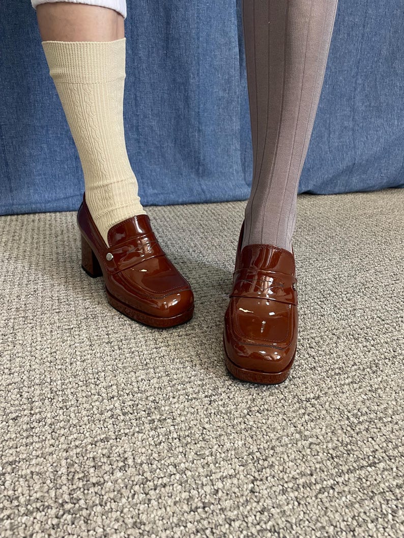 May include: Pair of brown patent leather platform loafers. The shoes have a square toe, a chunky heel, and a decorative strap across the vamp. One foot is wearing a cream-colored sock, and the other is wearing a gray ribbed stocking.