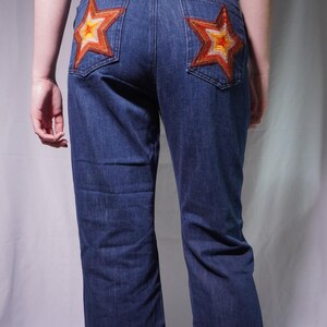 May include: Dark blue denim jeans featuring two back pockets, each embroidered with a multi-colored star design in shades of orange, yellow, and brown. The jeans have a standard waistband with belt loops and a straight-leg cut.