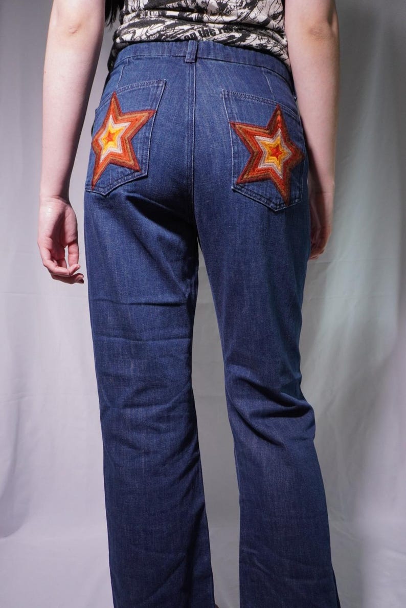 May include: Dark blue denim jeans featuring two back pockets, each embroidered with a multi-colored star design in shades of orange, yellow, and brown. The jeans have a standard waistband with belt loops and a straight-leg cut.