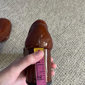 May include: A brown patent leather shoe with a block heel. The heel is approximately 2.5 inches tall, as measured with a pink measuring tape. The shoe is glossy and appears to be new.