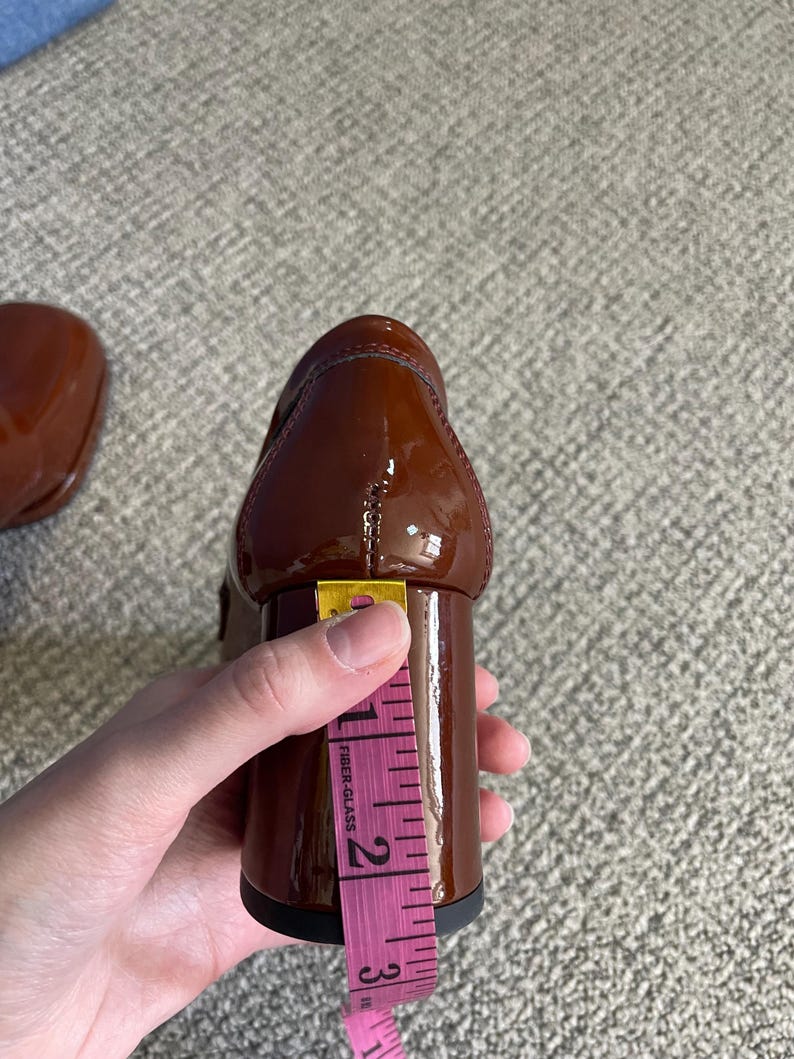 May include: A brown patent leather shoe with a block heel. The heel is approximately 2.5 inches tall, as measured with a pink measuring tape. The shoe is glossy and appears to be new.