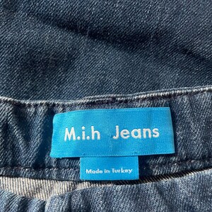 May include: Dark blue denim jeans featuring a bright blue rectangular fabric label stitched at the waistband. The label displays the white text M.i.h Jeans and the smaller text Made in Turkey. A dark brown button is visible below the label.