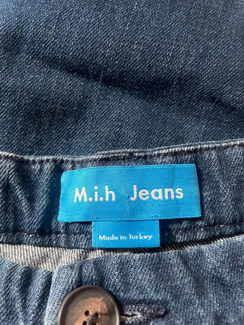 May include: Dark blue denim jeans featuring a bright blue rectangular fabric label stitched at the waistband. The label displays the white text M.i.h Jeans and the smaller text Made in Turkey. A dark brown button is visible below the label.