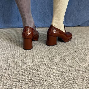 May include: A pair of brown patent leather heeled loafers. The shoes have a chunky heel and a rounded toe. One leg is wearing a pair of ribbed gray socks, and the other is wearing cream-colored cable knit socks. The shoes are on a gray carpet.