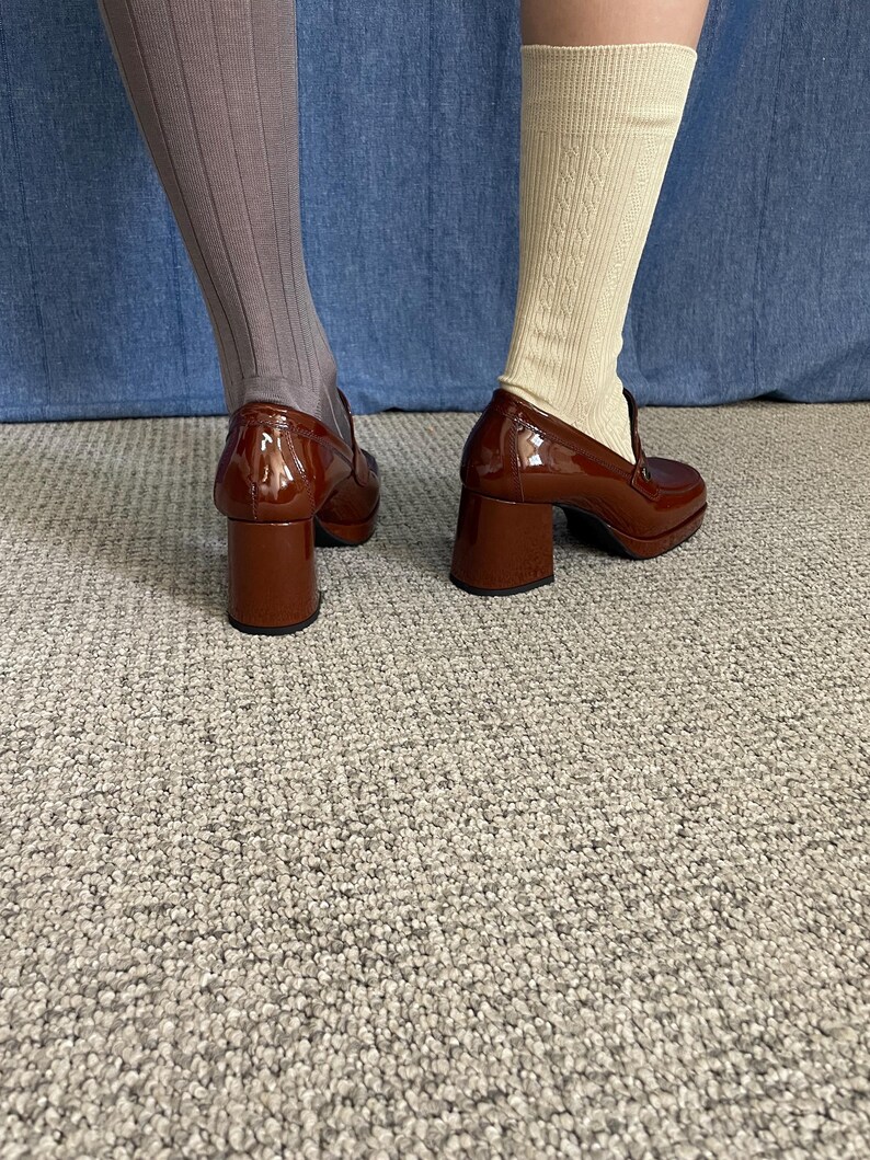 May include: A pair of brown patent leather heeled loafers. The shoes have a chunky heel and a rounded toe. One leg is wearing a pair of ribbed gray socks, and the other is wearing cream-colored cable knit socks. The shoes are on a gray carpet.