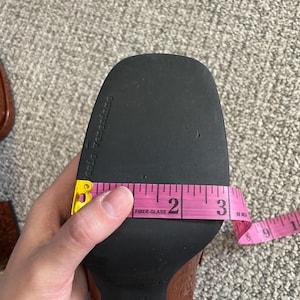 May include: A brown patent leather shoe sole is measured with a pink measuring tape. The sole is approximately 3 inches (7.6 cm) wide. The shoe is a size 37.