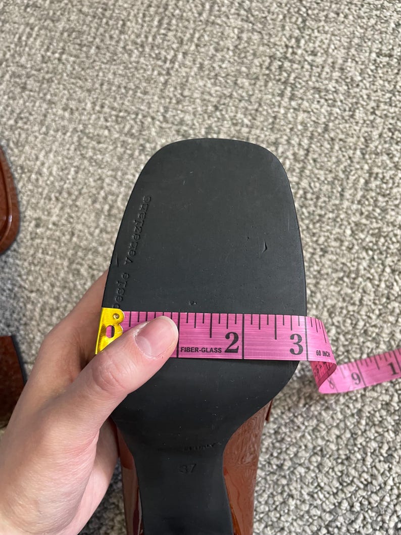 May include: A brown patent leather shoe sole is measured with a pink measuring tape. The sole is approximately 3 inches (7.6 cm) wide. The shoe is a size 37.