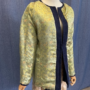May include: A long-sleeved jacket featuring a light green fabric with a dense, intricate floral pattern in shades of yellow and orange. The garment has a dark blue trim along the open front, neckline, and cuffs, and includes a single button closure.