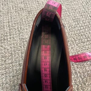 May include: A close-up of a shiny, burgundy shoe with a pink measuring tape inside. The tape shows measurements from 11 to 31, indicating the shoe's internal length. The shoe's glossy surface reflects light, highlighting its shape and design.