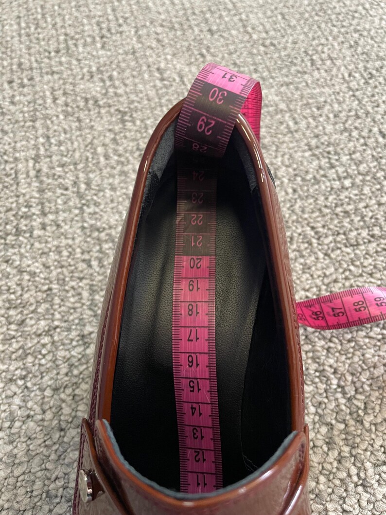May include: A close-up of a shiny, burgundy shoe with a pink measuring tape inside. The tape shows measurements from 11 to 31, indicating the shoe's internal length. The shoe's glossy surface reflects light, highlighting its shape and design.