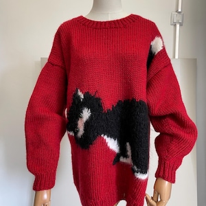 Pode incluir: Uma camisola de malha vermelha brilhante de manga comprida com um gráfico grande e felpudo de um gato preto e branco na frente. A camisola tem um decote redondo canelado, punhos canelados e uma bainha canelada, feita com fio grosso e texturizado.