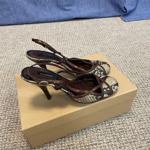 May include: A pair of brown and snakeskin-patterned high-heeled shoes with slingback straps. The shoes are on a cardboard box. The heels are approximately 7.6 cm tall.