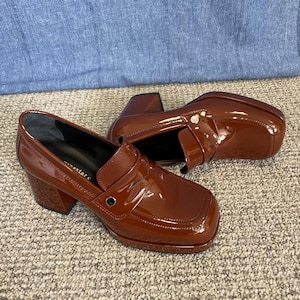 May include: A pair of glossy, brown leather heeled loafers. The shoes have a square toe, a strap across the top, and a chunky heel. The shoes are sitting on a neutral-colored carpet.
