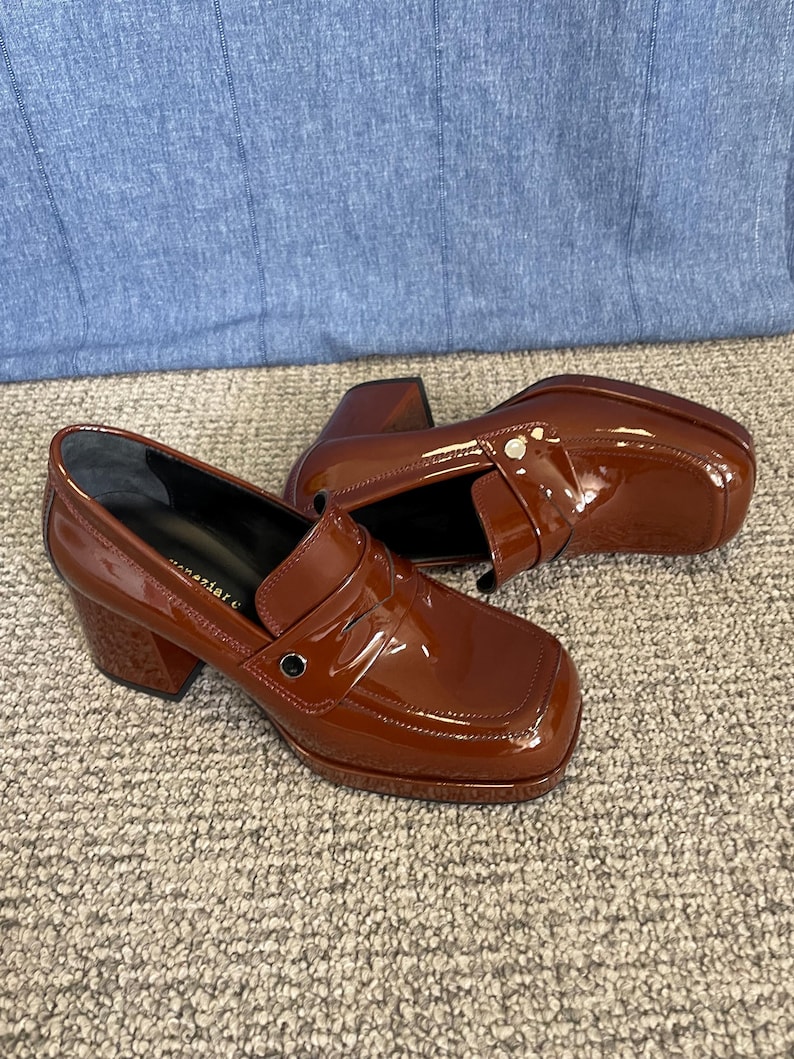 May include: A pair of glossy, brown leather heeled loafers. The shoes have a square toe, a strap across the top, and a chunky heel. The shoes are sitting on a neutral-colored carpet.