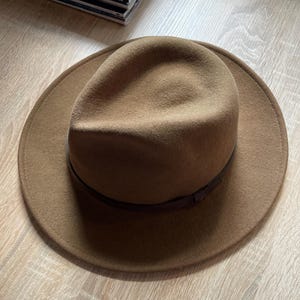 May include: A tan felt fedora hat with a wide brim and a dark brown band encircling the base of the crown. The hat features a pinched crown design and a smooth, uniform texture throughout the felt material, presenting a classic structured shape.