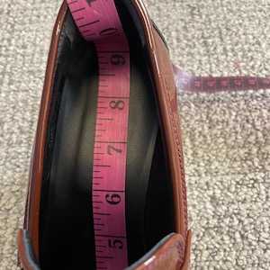 May include: A shiny, reddish-brown shoe with a black interior is being measured with a pink measuring tape. The tape shows a measurement of approximately 6 inches. The shoe has a low heel and a decorative silver accent.