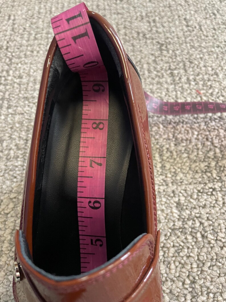 May include: A shiny, reddish-brown shoe with a black interior is being measured with a pink measuring tape. The tape shows a measurement of approximately 6 inches. The shoe has a low heel and a decorative silver accent.