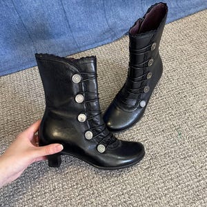 Könnte beinhalten: Ein Paar schwarze Leder-Ankle Boots mit dekorativen silbernen Knöpfen und Spitzenbesatz. Die Stiefel haben einen kleinen Absatz und sind vorne geschnürt. Die Stiefel werden auf einem neutralen Teppich präsentiert.