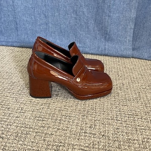 May include: A pair of brown patent leather heeled loafers. The shoes have a square toe, a small gold accent, and a block heel. The shoes are sitting on a neutral-colored carpet.