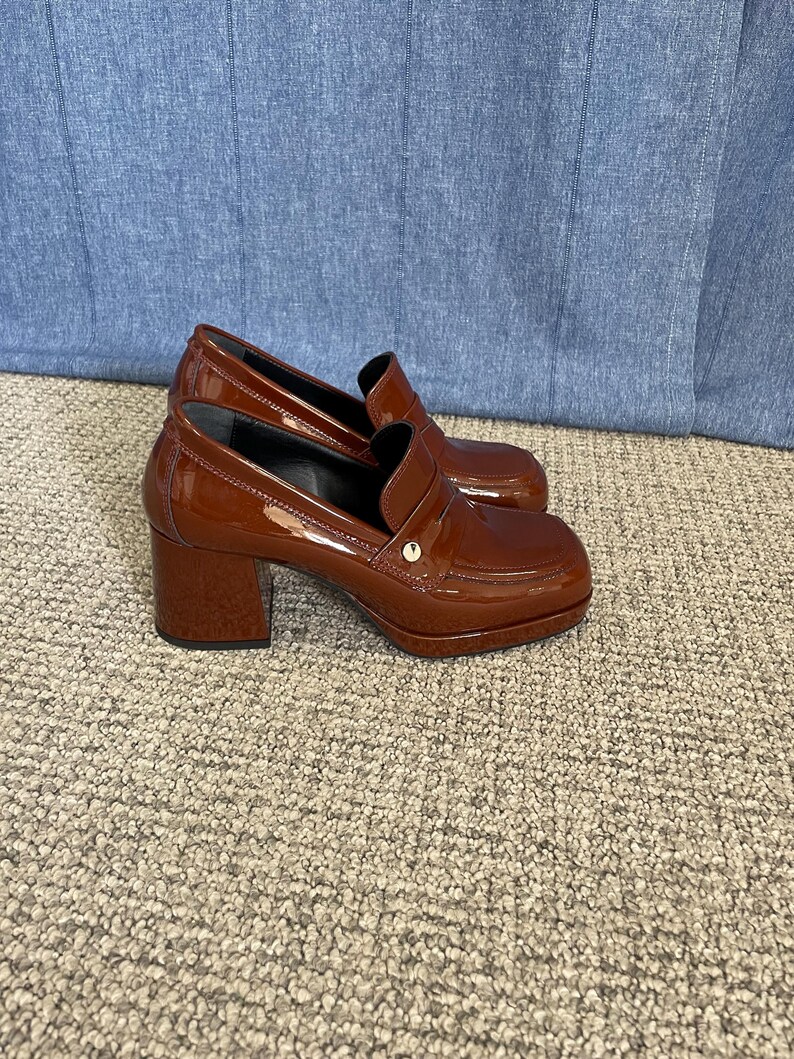 May include: A pair of brown patent leather heeled loafers. The shoes have a square toe, a small gold accent, and a block heel. The shoes are sitting on a neutral-colored carpet.
