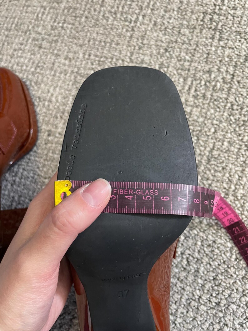 May include: Close-up of a brown patent leather shoe with a black sole. A pink measuring tape shows the shoe's width. The shoe size is 37. The sole is marked "pesie Veneziane".