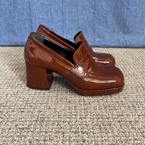 May include: A pair of brown patent leather heeled loafers. The shoes have a square toe, a chunky heel, and a small silver button detail on the vamp. The heels measure approximately 3 inches.