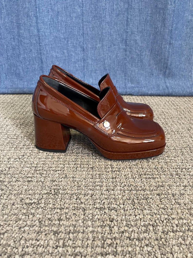 May include: A pair of brown patent leather heeled loafers. The shoes have a square toe, a chunky heel, and a small silver button detail on the vamp. The heels measure approximately 3 inches.