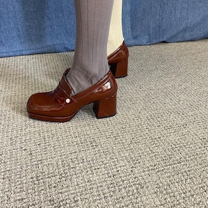 May include: Brown patent leather platform shoes with a chunky heel. The shoes have a rounded toe and a strap across the top. The shoes are worn with ribbed gray socks and a pair of beige socks. The shoes are approximately 3 inches (7.6 cm) high.