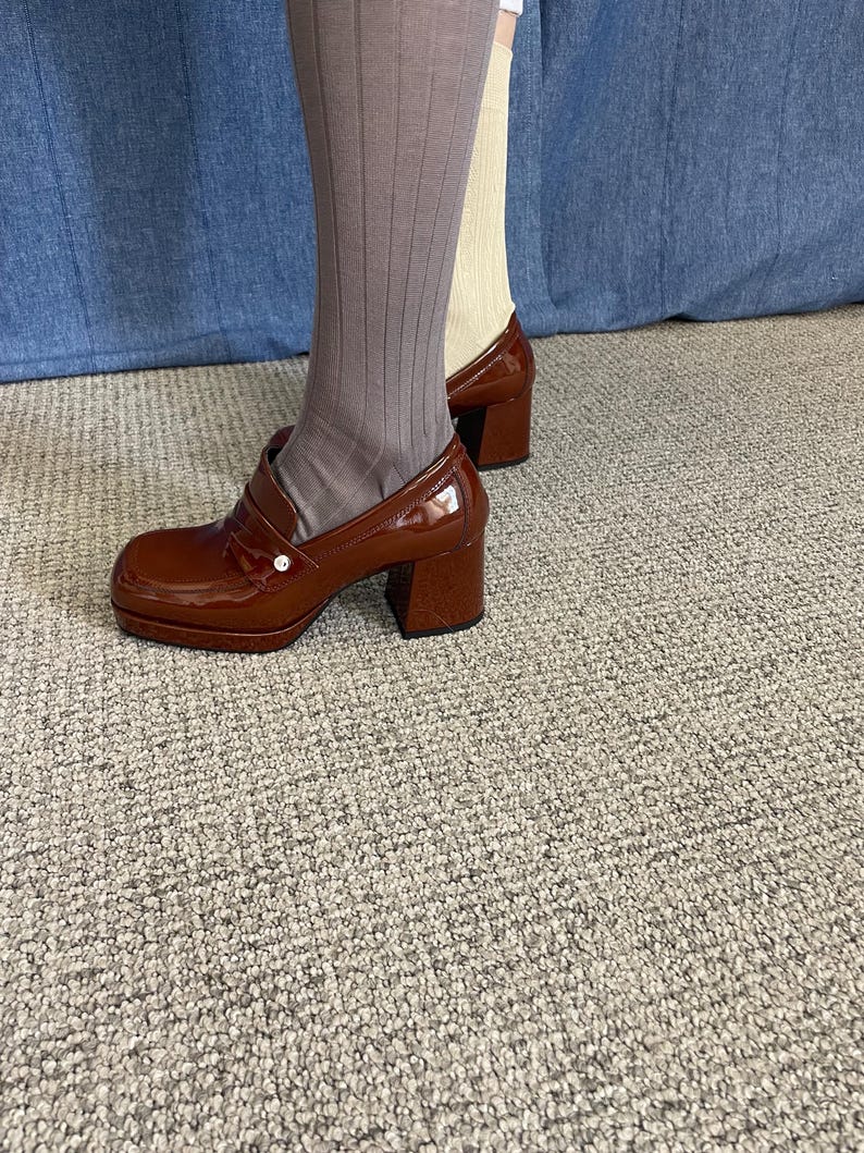 May include: Brown patent leather platform shoes with a chunky heel. The shoes have a rounded toe and a strap across the top. The shoes are worn with ribbed gray socks and a pair of beige socks. The shoes are approximately 3 inches (7.6 cm) high.