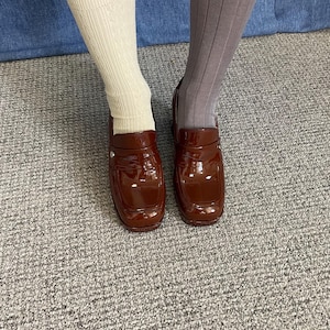 May include: A pair of glossy, brown loafers with a square toe. The shoes are worn with a cream-colored sock on one leg and a gray sock on the other. The background is a denim material and a gray carpet.