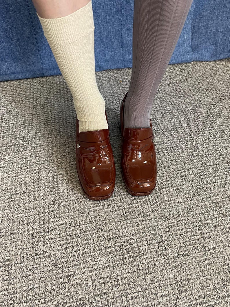 May include: A pair of glossy, brown loafers with a square toe. The shoes are worn with a cream-colored sock on one leg and a gray sock on the other. The background is a denim material and a gray carpet.