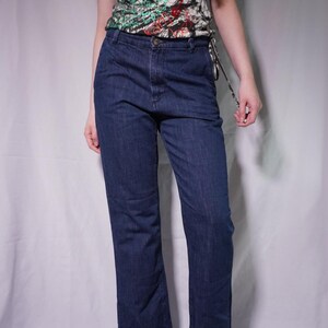 May include: Dark wash denim jeans with a straight leg cut. The jeans have a button closure and a classic five-pocket design. The model is wearing a patterned tank top with red, green, and black accents. The shoes are dark red flats.