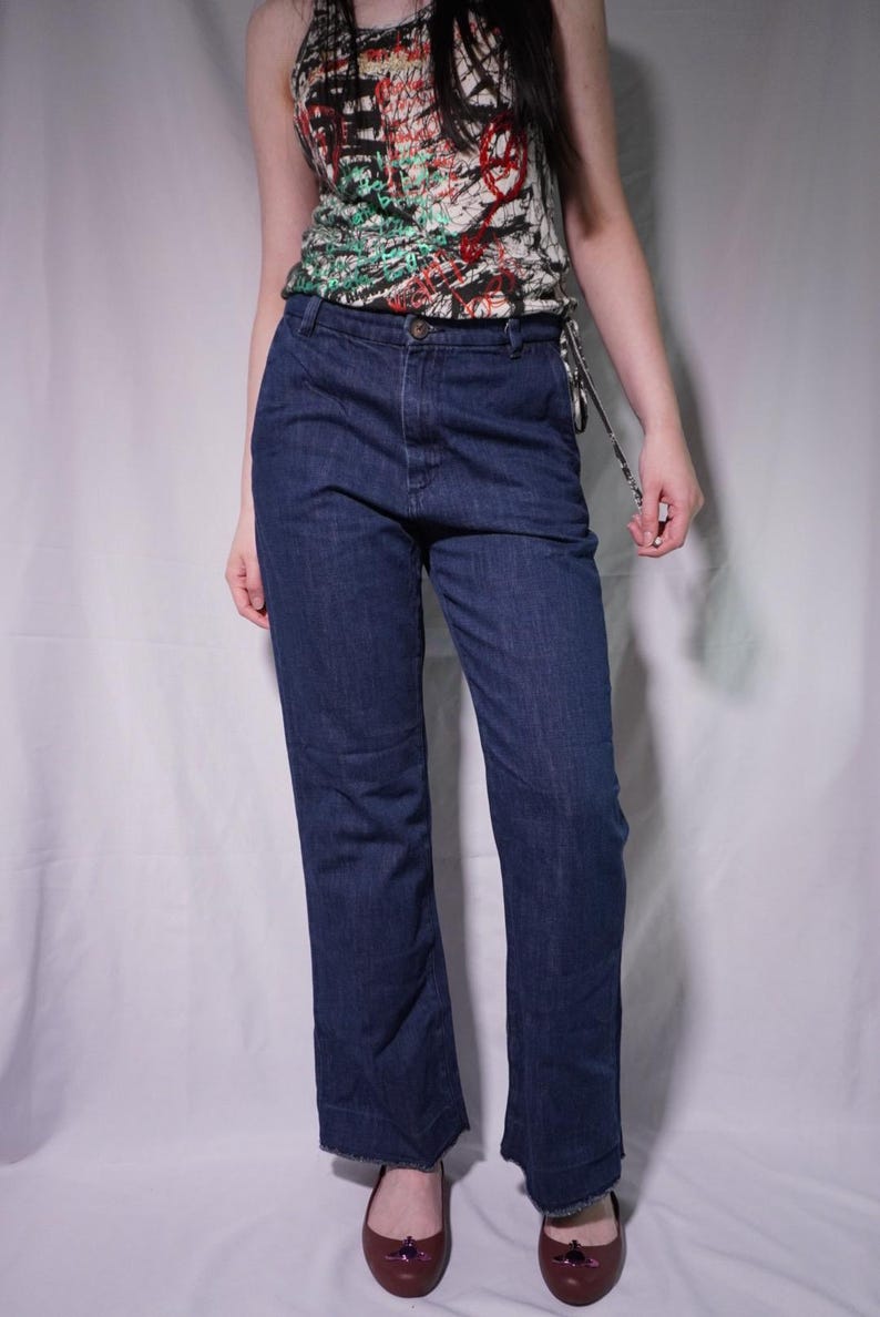 May include: Dark wash denim jeans with a straight leg cut. The jeans have a button closure and a classic five-pocket design. The model is wearing a patterned tank top with red, green, and black accents. The shoes are dark red flats.