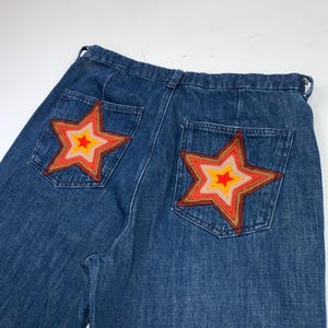 May include: Blue denim jeans featuring two back pockets, each adorned with a large, multi-layered star patch. The star patches consist of concentric rings in shades of red, orange, yellow, and light pink, stitched onto the denim fabric.