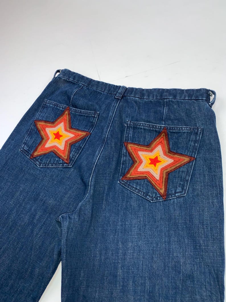 May include: Blue denim jeans featuring two back pockets, each adorned with a large, multi-layered star patch. The star patches consist of concentric rings in shades of red, orange, yellow, and light pink, stitched onto the denim fabric.