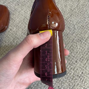 May include: A close-up of a brown patent leather shoe with a block heel. A measuring tape shows the heel height is approximately 3 inches. The shoe has a glossy finish and a classic design.
