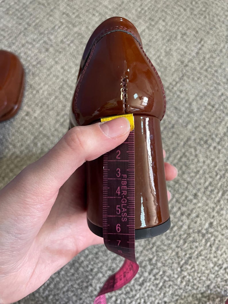 May include: A close-up of a brown patent leather shoe with a block heel. A measuring tape shows the heel height is approximately 3 inches. The shoe has a glossy finish and a classic design.