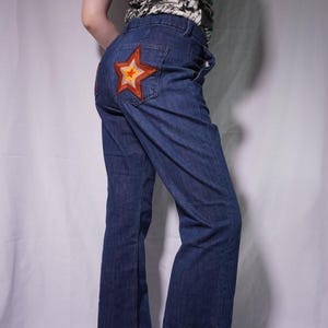 May include: Dark blue denim jeans with a large, embroidered star on the back pocket. The star features a red, orange, and yellow color scheme. The jeans have a flared leg design.
