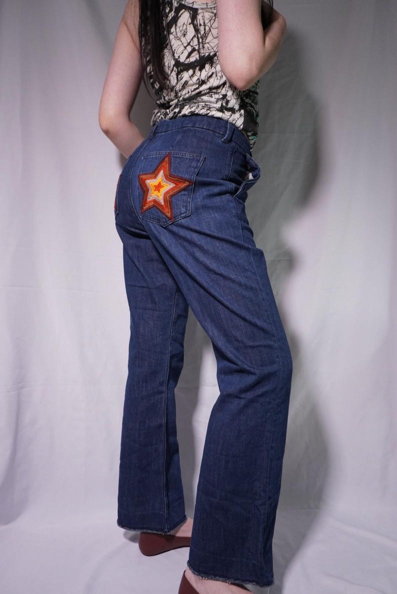 May include: Dark blue denim jeans with a large, embroidered star on the back pocket. The star features a red, orange, and yellow color scheme. The jeans have a flared leg design.