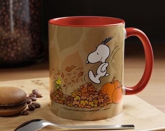 May include: A ceramic mug with a bright orange interior and handle, featuring a graphic of Snoopy jumping into a pile of autumn leaves with Woodstock nearby and two orange pumpkins. The mug has a tan background with a subtle leaf pattern.
