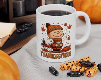 May include: White ceramic mug featuring Charlie Brown and Snoopy in fall attire, holding a cup of coffee. The mug has the word "THANKSGIVING" in orange. Autumn leaves and pumpkins surround the characters, perfect for the Thanksgiving holiday.