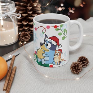 Puede incluir: Taza de cerámica blanca con los personajes de Bluey y Bingo con gorros de Papá Noel, sosteniendo una galleta, con decoraciones navideñas. La taza tiene un asa curva y está llena de un líquido oscuro.