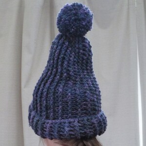 Pode incluir: Um gorro de malha roxo e azul com um pompom felpudo no topo.