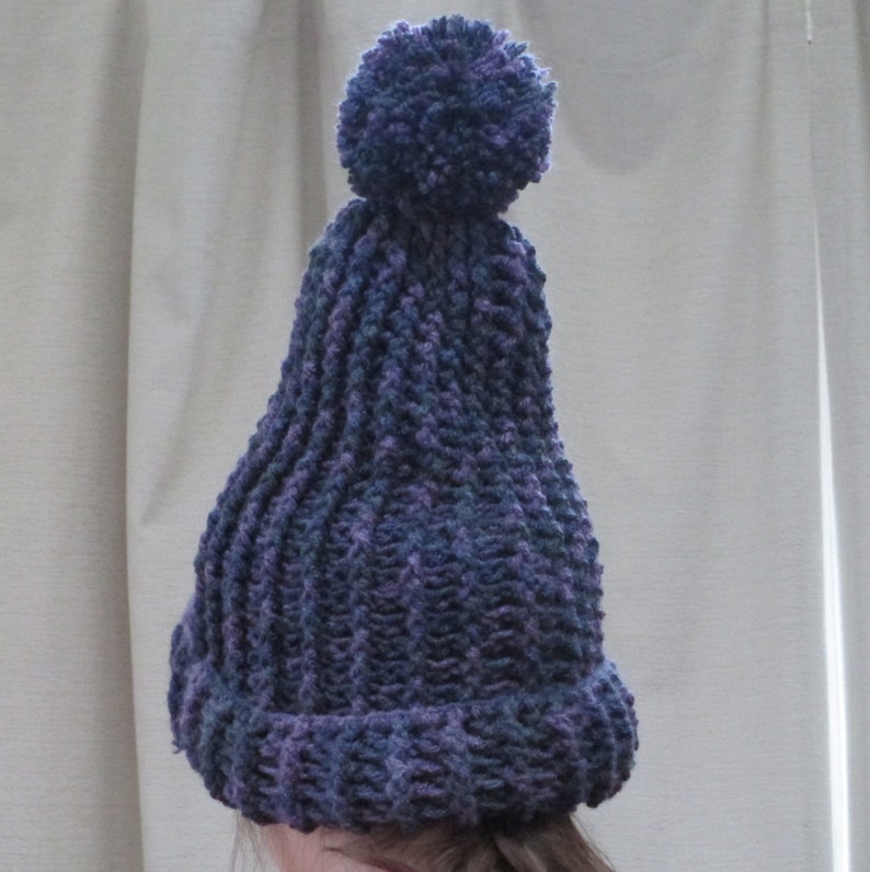 Pode incluir: Um gorro de malha roxo e azul com um pompom felpudo no topo.