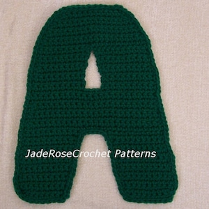 May include: A green crocheted letter A. The letter is made with a single crochet stitch and has a slightly rounded shape. The letter is on a white background.