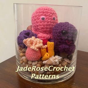 May include: A clear glass vase filled with decorative crochet sea creatures, including a pink octopus, a purple octopus, and coral. The vase sits on a bed of small stones and seashells. The text "JadeRoseCrochet Patterns" is visible on the front of the vase.